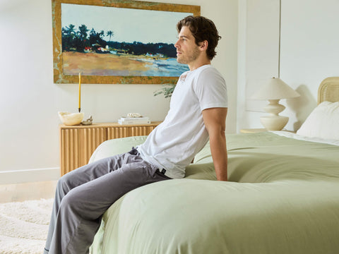 A young man sitting at the end of his neatly made bed with Avec linen blend bedding